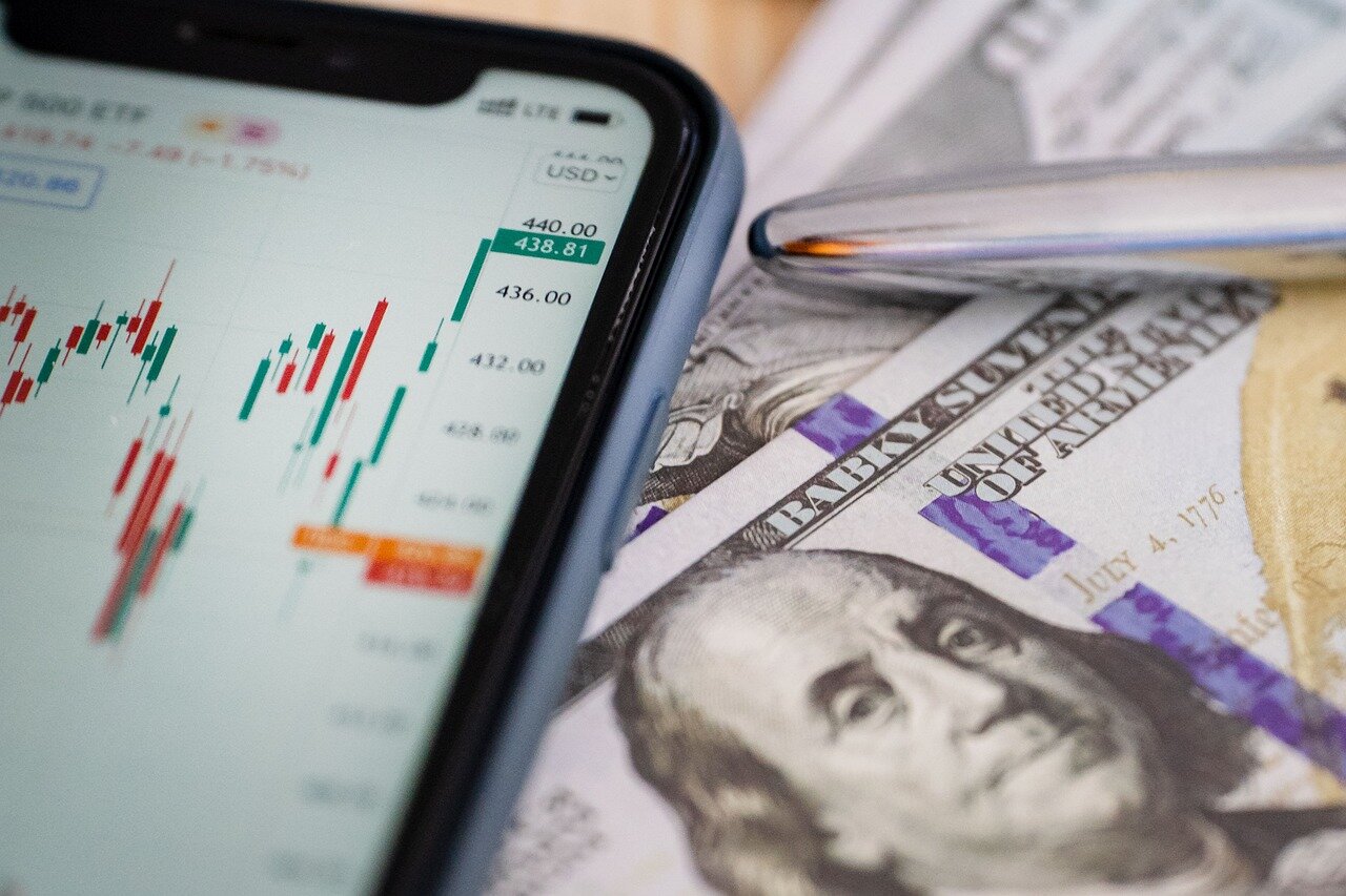 A diverse group of young adults gathers around a table, discussing investment strategies and reviewing financial apps on their smartphones in a modern coworking space.
