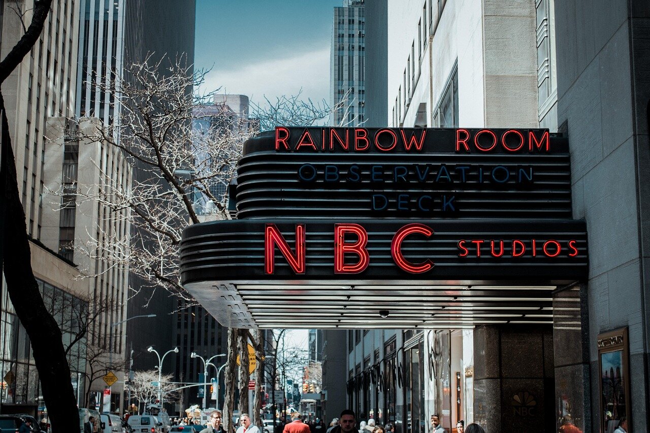 Media executives gather in a conference room, discussing NBC's historical impact on television broadcasting while referencing the evolution of reality TV and its cultural significance.