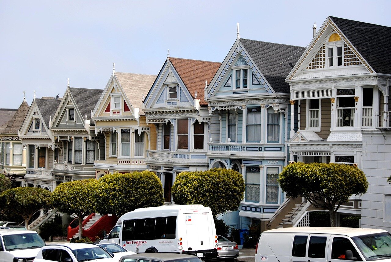 A bustling San Francisco street showcases a row of colorful homes with "For Sale" signs, reflecting the city's booming real estate market amid rising demand from the tech industry.